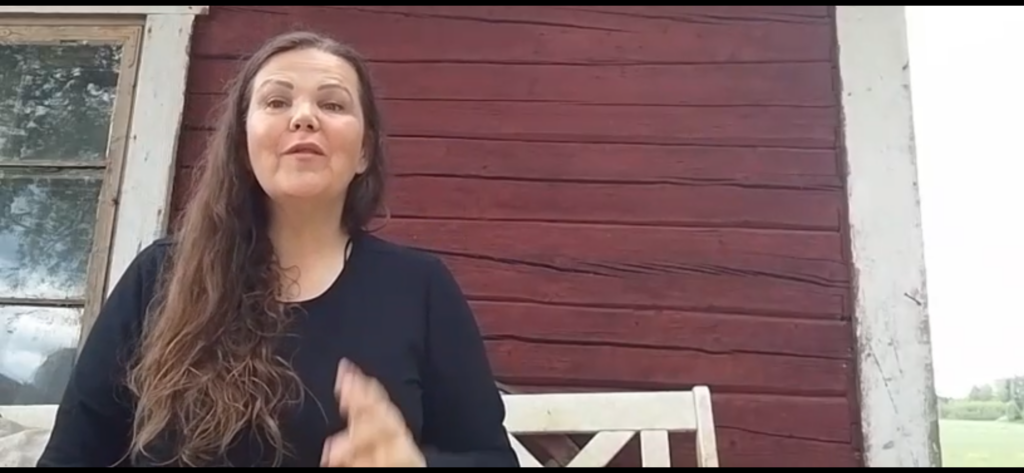 Anne istuu valkoisella puupenkillä punaisen hirsitalon edustalla.
